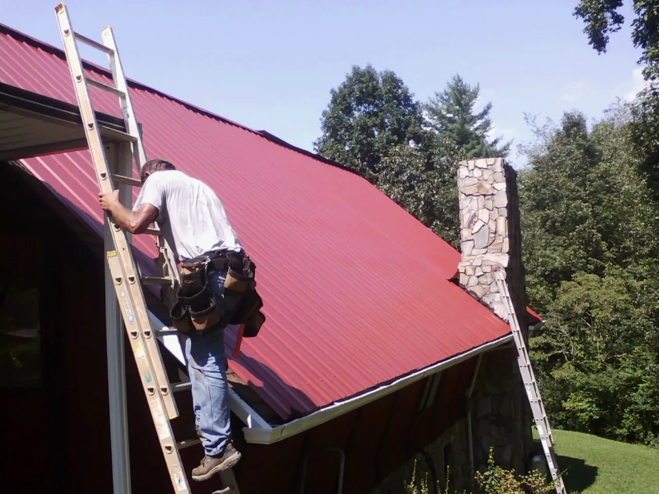 Roofer on a ladder beside a red metal roof and stone chimney — Soffit & Fascia Repair by Roofing Brothers in Pauline